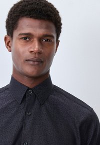 Schwarzes Hemd mit Muster und Knopfkragen, das ein dezentes Design auf einem strukturierten Stoff zeigt. Nahaufnahme eines Porträts einer Person, die es trägt.