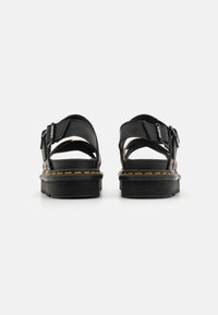 Sort svarte sandaler med justerbare stropper, en tykk svart såle og gule stikningsdetaljer langs kanten. Åpen bakdesign med teksturert overflate.