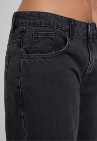 Jean en denim noir porté à la taille, montrant les poches avant et latérales ainsi que les passants de ceinture sur une personne avec la peau visible au-dessus de la ceinture.