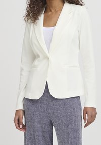 Bílý blazer s přiléhavým střihem, dlouhými rukávy, s klopami a bočními kapsami. Kombinován s kalhotami v šikmém vzoru.