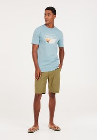 Camiseta de algodón azul claro con un diseño gráfico geométrico en naranja, verde y blanco. Combinada con pantalones cortos verdes oliva y chanclas.
