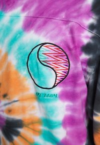 Koszulka tie-dye w różnych kolorach, z okrągłym haftowanym wzorem z falą i tekstem poniżej. Miękka faktura materiału.