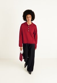 Pulôver de malha vermelho com colarinho e três botões, combinado com calças pretas soltas. Os acessórios incluem uma pequena bolsa vermelha texturizada e sapatos estampados.