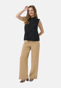 Črna čipkasta brezrokavnik s valovitimi robovi, v kombinaciji s svetlo bež širokimi hlačami in salonarji s leopardjim potiskom.