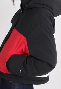 Černá a červená péřová bunda s kapucí, s barevným blokovým designem a elastickými manžetami. Obsahuje malé logo na rukávu.