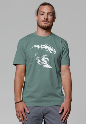 Groene katoenen T-shirt met een witte astronaut die surft, omgeven door een gestructureerd golfontwerp. Korte mouwen en ronde hals.
