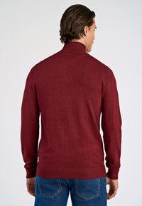 Burgunderfarbener Pullover mit hohem Kragen, gerippten Bündchen und Saum, aus glattem Strickstoff, getragen über blauen Jeans.
