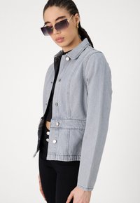 Veste en denim gris clair au design structuré, avec des boutons-pression et un col. Associée à un top noir court et un pantalon noir.