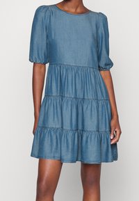 Vestido de mezclilla con volantes y mangas abullonadas, cuello redondo y secciones fruncidas. Color azul suave, tela ligera y silueta fluida.