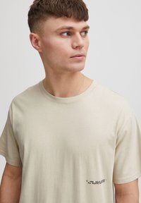 Beige bomulds t-shirt med korte ærmer, rund halsudskæring, med et lille sort tryk på venstre brystområde. Glat tekstur.