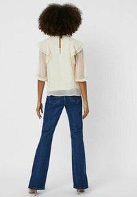 Cremefarvet bluse med rynkede ærmer og prikmønster, parret med bredde blå jeans. Bageste knapper lukning detaljer synlige.