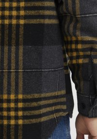 Flannel skjorte med et sort og gult ternet mønster, kontrasterende teksturer, knapmanchet og et afrundet hæmdesign.