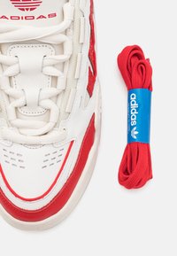 White sneaker with red accents, textured panels, and prominent lacing system. Includes a red shoelace with a blue Adidas label.