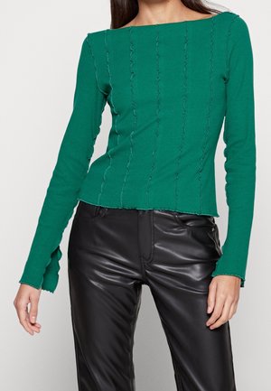 Mujer con una blusa verde de manga larga con costuras verticales con volantes y pantalones de cuero negros con bolsillos delanteros.