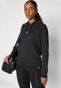 Moleton desportivo preto com um bolso frontal, apresentando um capuz com cordão e um logótipo de três riscas brancas. Usado com leggings pretas a condizer e uma mala.