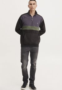 Sudadera de media cremallera en negro, púrpura y verde. Material de forro polar, con un bolsillo frontal y un ajuste ergonómico. Combinada con unos vaqueros oscuros y zapatillas.