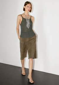 Top in maglia grigia con spalline sottili, abbinato a culottes in suede verde oliva con uno spacco laterale. Una collana con pendente argento aggiunge brillantezza.