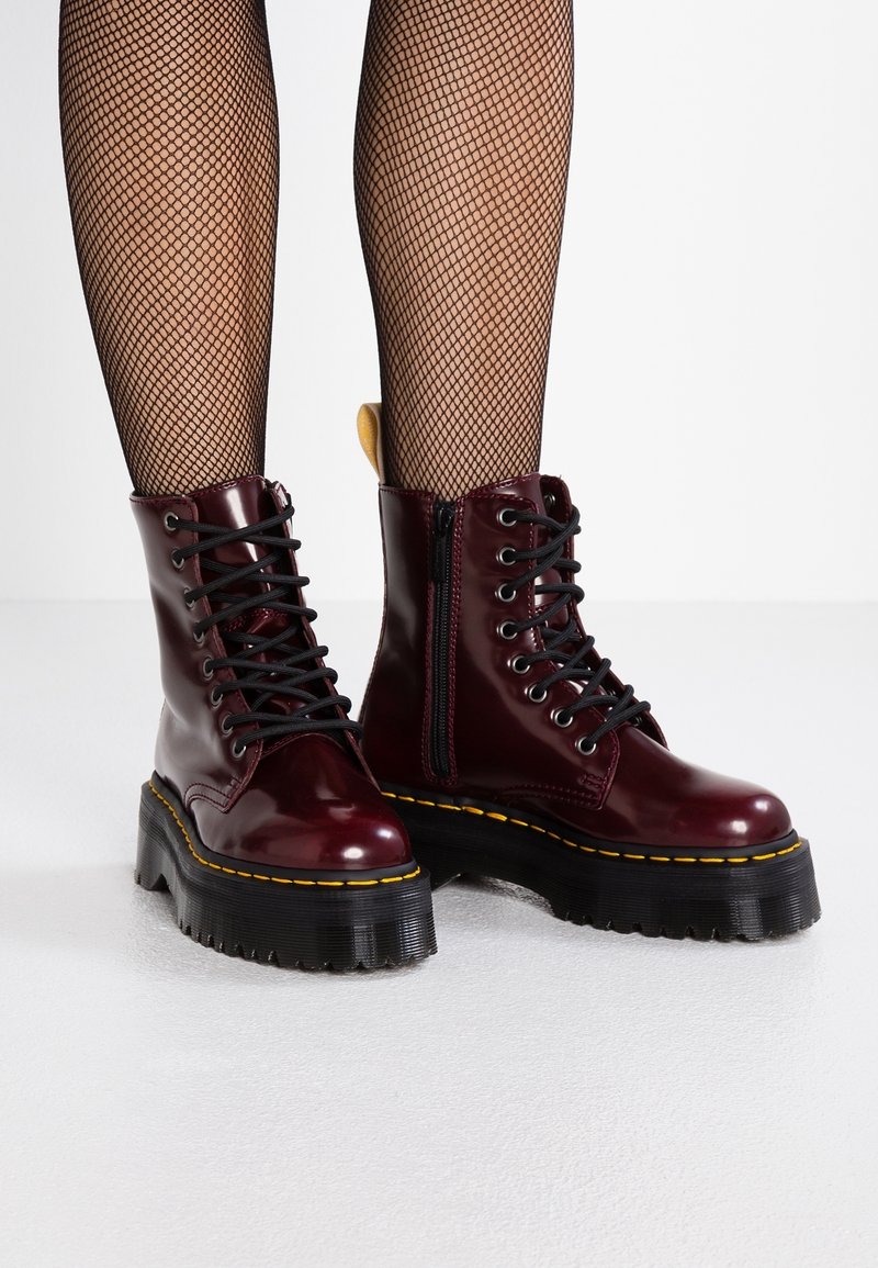 Bottes à plateforme bordeaux brillantes de style combat avec lacets noirs, semelles épaisses à surpiqûres jaunes, portées avec des collants résille noirs sur un sol blanc.