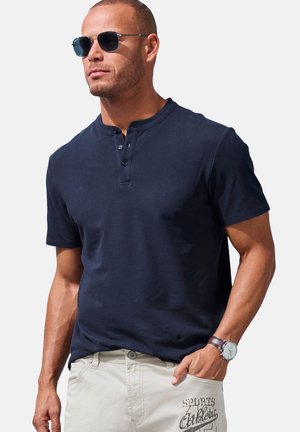 Dunkelblaues Baumwoll-Polohemd mit kurzen Ärmeln, einer Knopfleiste mit drei Knöpfen und rundem Halsausschnitt. Getragen mit hellen Hosen und einer Uhr.