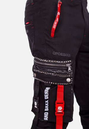 Black cargo pants with multiple pockets, red accents, silver studs, and textured zippers. Features brand logo and adjustable straps.