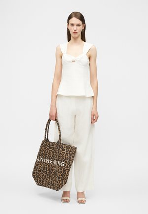 Femeie îmbrăcată cu un top alb fără mâneci și pantaloni largi, ținând o geantă mare de umăr cu imprimeu leopard și text alb, în picioare pe un fundal gri simplu.