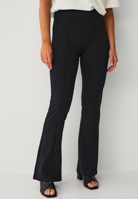 Sorte flared leggings med høj talje, lavet af strækbart materiale. Har sømdetaljer langs benene. Matchet med sorte blokhælede sandaler.