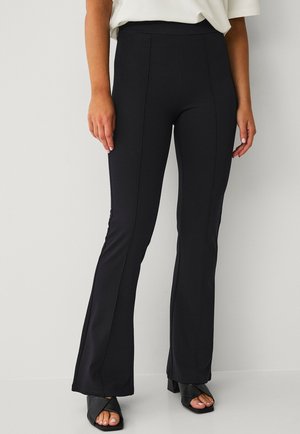 Zwarte flare leggings met een hoge tailleband, gemaakt van rekbaar materiaal. Heeft naaddetails langs de benen. Gecombineerd met zwarte sandalen met blokhak.