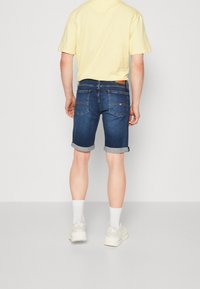 Mörkblå denimshorts med uppvikta muddar, kombinerade med en gul t-shirt. Baksidan har två fickor och en brun lädertag.
