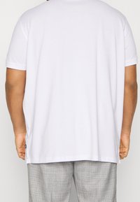 Mężczyzna w gładkiej białej koszuli z krótkim rękawem i szarych kratkowanych spodniach, pokazany z tyłu na jednolitym tle.