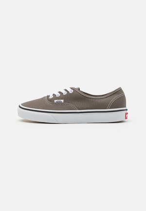 Zapatillas de lona marrón con cordones y costuras blancas, que cuentan con una suela de goma blanca y un pequeño detalle del logo rojo en el talón.
