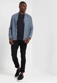 Lys blå knapp-up skjorte over en marineblå T-skjorte med en blomsterlomme, kombinert med svarte skinny jeans og svarte sneakers.