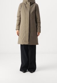 Pudset olivengrøn jakke med høj krave, lynlås foran og to sidelommer, kombineret med brede sorte bukser og sorte sko.