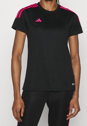 Kvinde iført sort Adidas sportstrøje med pink striber på skuldrene og pink Adidas-logo, kombineret med sorte bukser med pink detaljer.