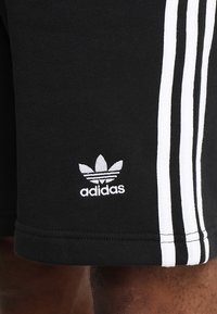 Pantaloni sportivi neri con una design a tre righe bianche lungo i lati. Presentano un logo Adidas ricamato bianco in basso a sinistra.