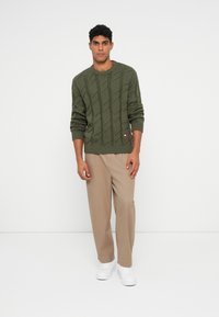 Olivegrüner Pullover mit Kabelmuster, gerippten Bündchen und Saum, kombiniert mit hellbeigen Hosen und weißen Sneakern.