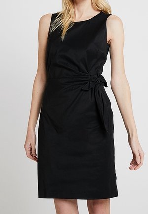 Kvinde iført ærmeløs sort knælængde kjole med rund halsudskæring og bindebånd i siden ved taljen mod en ensfarvet baggrund.