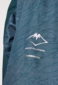 Teal løbejakke med et struktureret mønster af prikker og bølger. Har hvidt logo "ASICS FUJITRAIL" og reflekterende detaljer. Letvægtsmateriale.