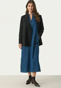 Černé přizpůsobené sako přes šaty z denimu po kolena s přední kapsou. Černé podpatky a punčochy s tečkami dotvářejí celý outfit.