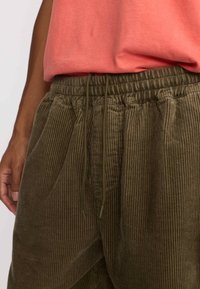 Olijfgroene corduroy broek met verticale strepen, elastische tailleband en verstelbare koord. Voorzien van zijzakken en een zachte textuur.