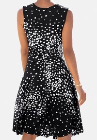 Kvinna i en ärmlös svart klänning med spridda vita prickar, sedd från baksidan, med guldsmycken på höger handled.