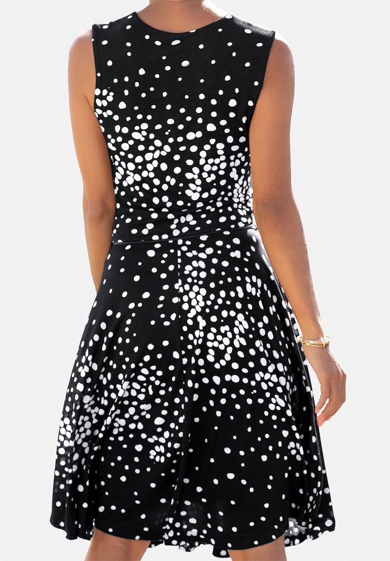 Kvinna i en ärmlös svart klänning med spridda vita prickar, sedd från baksidan, med guldsmycken på höger handled.