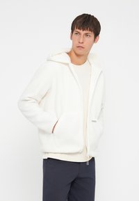 White fleece zip-up hoodie with a drawstring, front pockets, and a soft texture, worn over a light ribbed shirt and paired with dark pants.