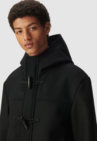 Cappotto nero con cappuccio, chiusure a soffietto, design strutturato e tessuto testurizzato. Presenta un colletto alto e cuciture sottili.