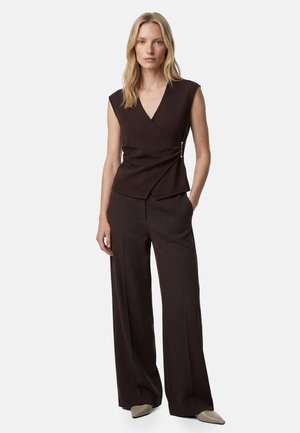 Vrouw staand, draagt een mouwloze zwarte wikkeltop met zijknopen en bijpassende wijde broek, beige puntige schoenen, neutrale achtergrond.