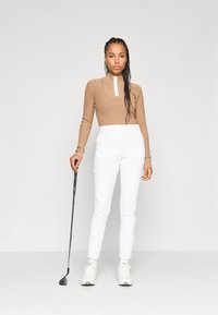 Brūns ribots krekls ar garām piedurknēm un ar rāvējslēdzēju, balti piegriezti bikses un balti sporta apavi. Labajā rokā tur golfu.