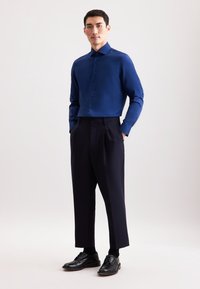 Man staat met zijn handen in zijn zakken, gekleed in een blauwe button-down shirt, zwarte cropped broeken, zwarte sokken en zwarte nette schoenen op een effen achtergrond.