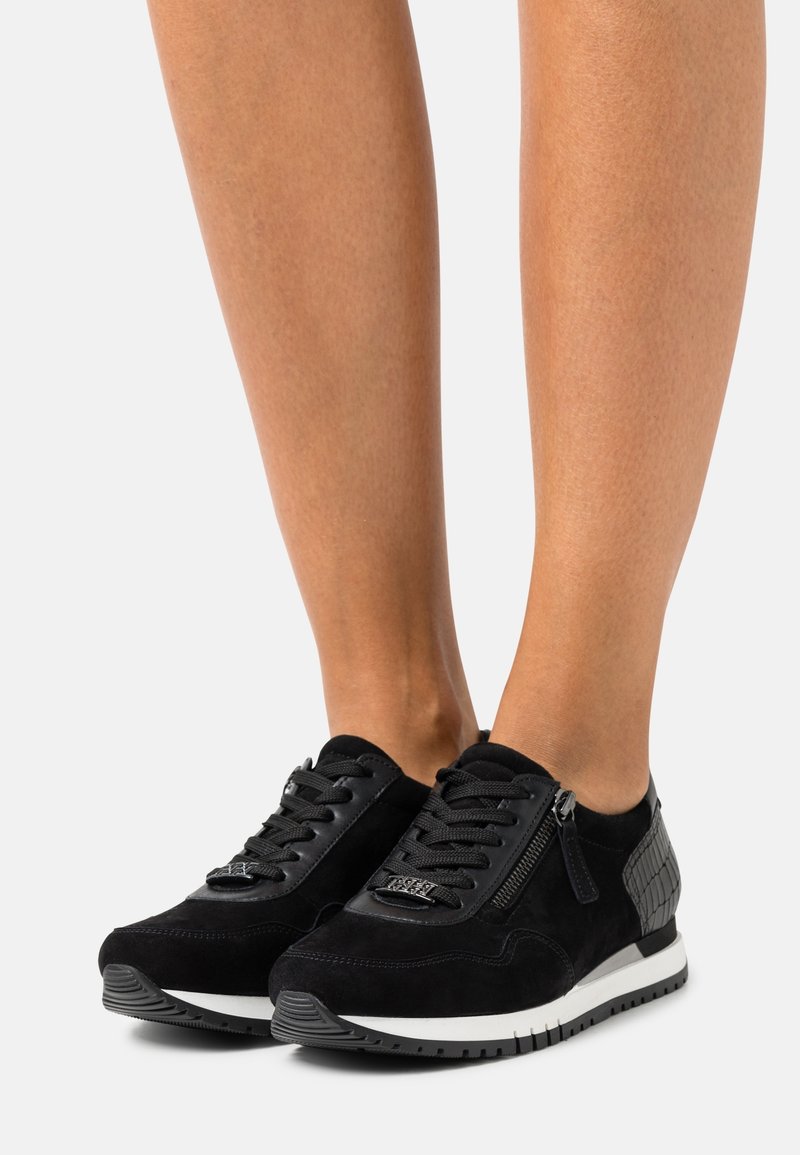 Jambes portant des baskets noires avec des fermetures éclair latérales, des panneaux texturés au talon et des semelles blanches, debout sur une surface blanche.