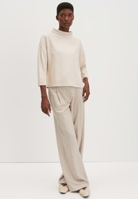 Top beige chiaro oversize con collo alto e maniche tre quarti, abbinato a pantaloni ampi plissettati beige chiaro e scarpe slip-on color crema.