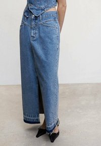 Falda maxi de denim en azul claro con una abertura frontal, dobladillo deshilachado y acentos en denim oscuro en contraste. Se lleva con zapatos negros de punta.