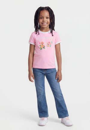 Pige med flettet hår iført en lyserød t-shirt med blomstermotiv og logo, blå jeans og hvide sneakers, stående foran en hvid baggrund.